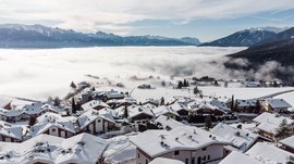 The mountain village of Meransen The picture shows a village with snow-covered houses on a hill, surrounded by a layer of fog. In the background, impressive snow-covered mountain peaks are visible under a partly cloudy sky.