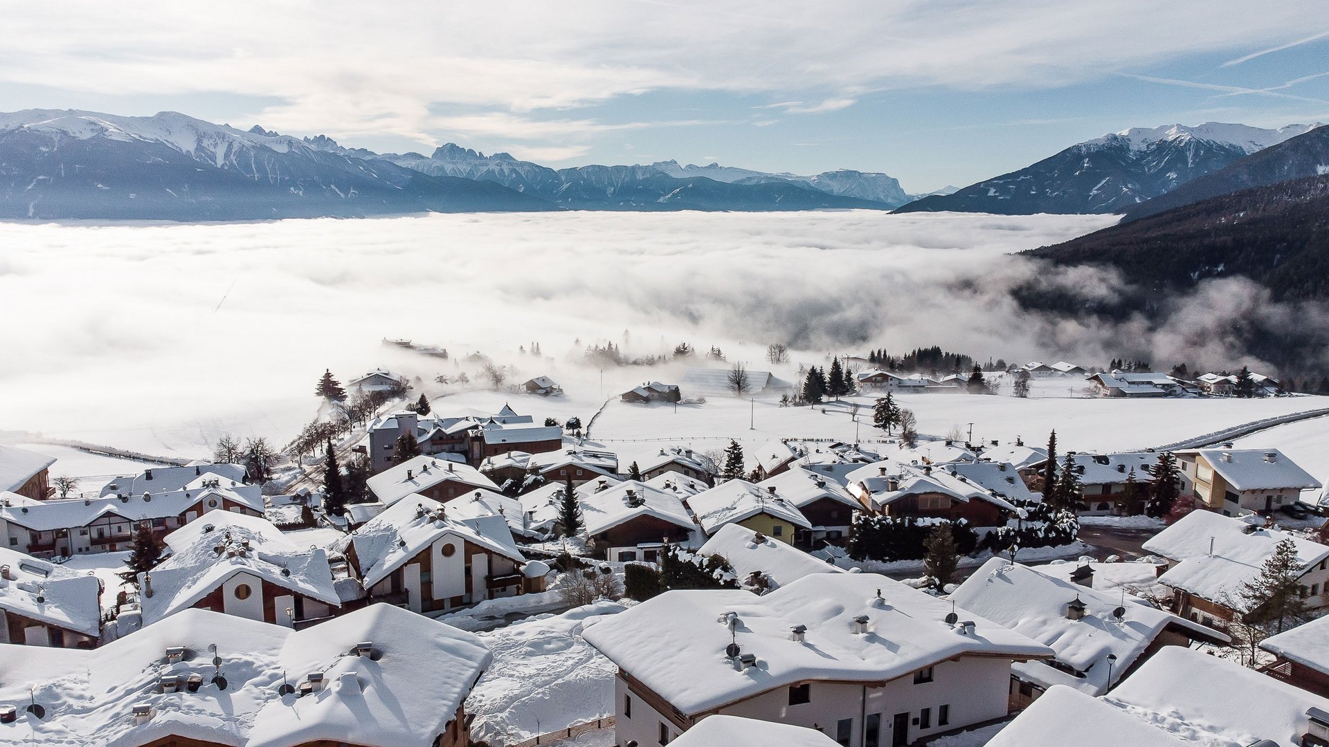 The mountain village of Meransen The picture shows a village with snow-covered houses on a hill, surrounded by a layer of fog. In the background, impressive snow-covered mountain peaks are visible under a partly cloudy sky.