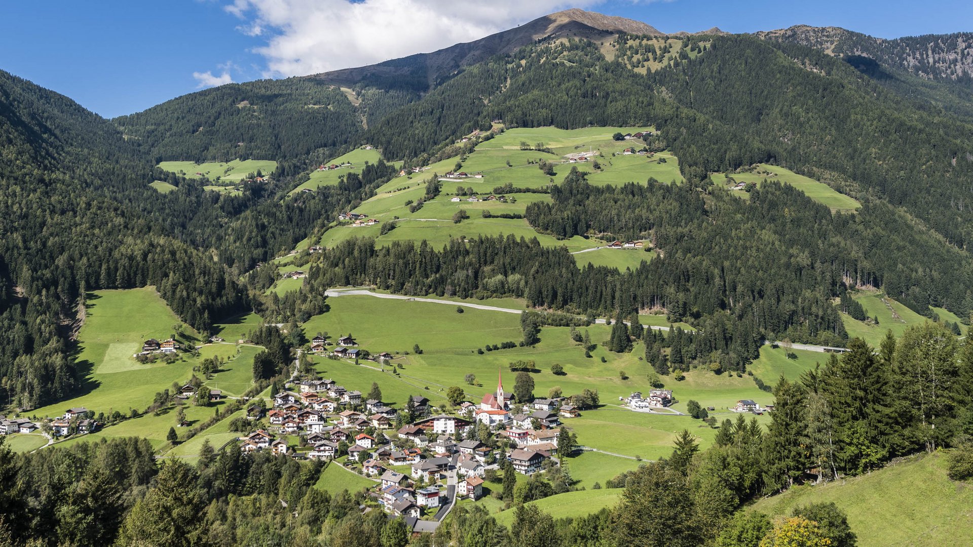 Pfunders: urig und idyllisch Das Bild zeigt eine grüne, hügelige Landschaft mit einem kleinen Dorf, das in einem Tal liegt. Die Umgebung ist von dichten Wäldern und Bergen umgeben, während der Himmel klar und blau ist, mit vereinzelten weißen Wolken.