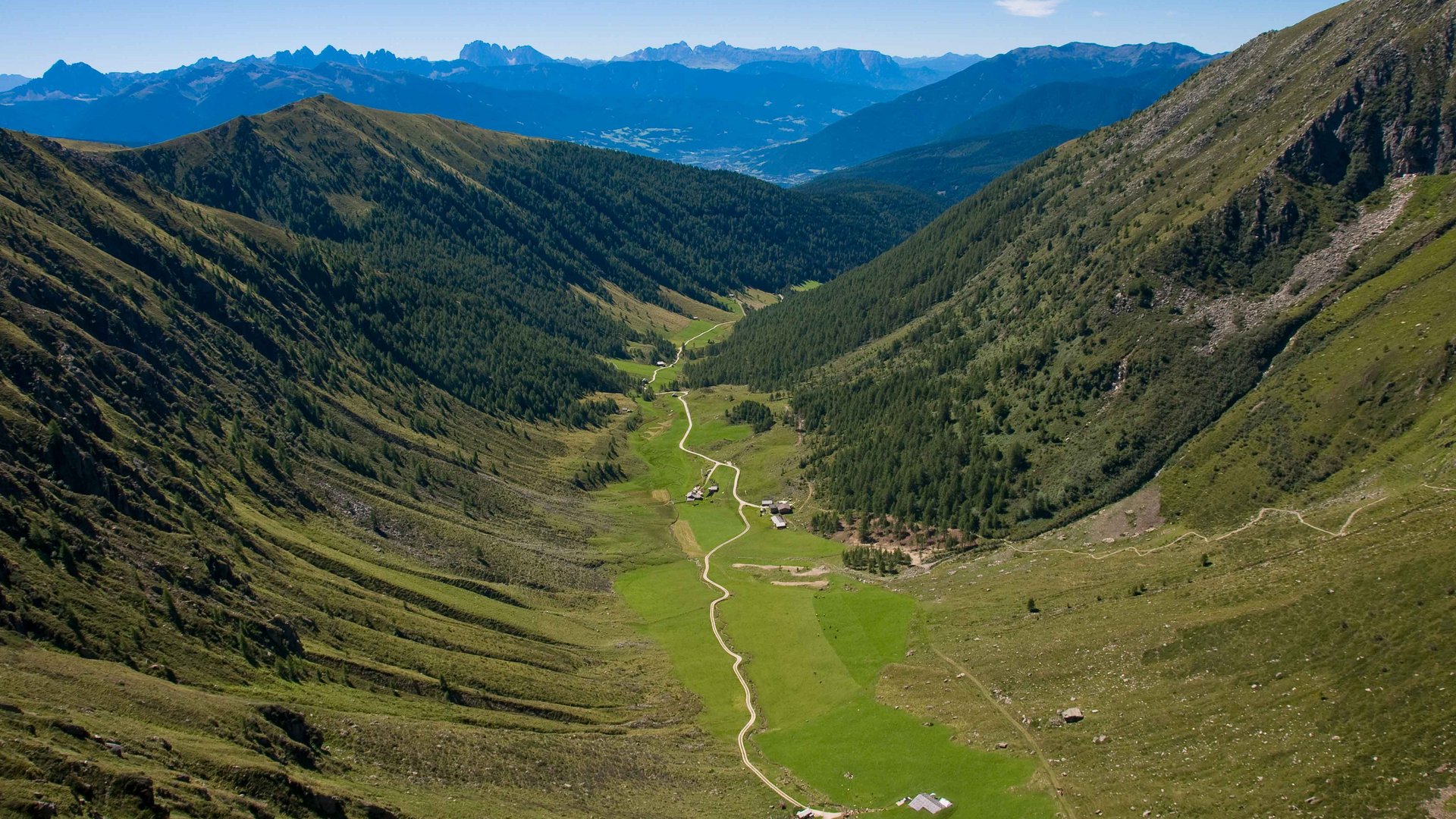 The Altfasstal valley, Meransen The picture shows a wide valley stretching between two tall, forested mountain slopes. A narrow, winding path leads through the green valley, past some scattered huts. In the background, the majestic peaks of the Alps can be seen dominating the horizon.