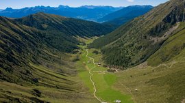 The Altfasstal valley, Meransen The picture shows a wide valley stretching between two tall, forested mountain slopes. A narrow, winding path leads through the green valley, past some scattered huts. In the background, the majestic peaks of the Alps can be seen dominating the horizon.