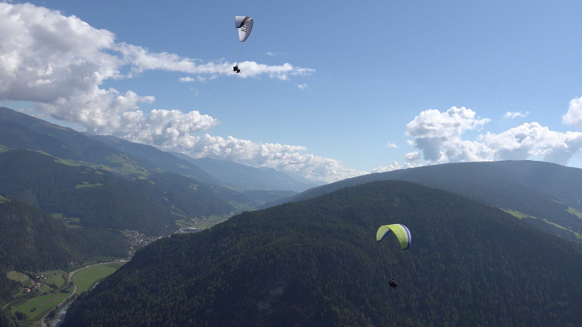 Paragliding v Jižním Tyrolsku Obrázek ukazuje dva paraglidingy, kteří létají nad rozsáhlou horskou krajinou s hustě zalesněnými kopci. Obloha je jasná s několika rozptýlenými mraky a v pozadí se rozkládají zelené údolí a další hory.
