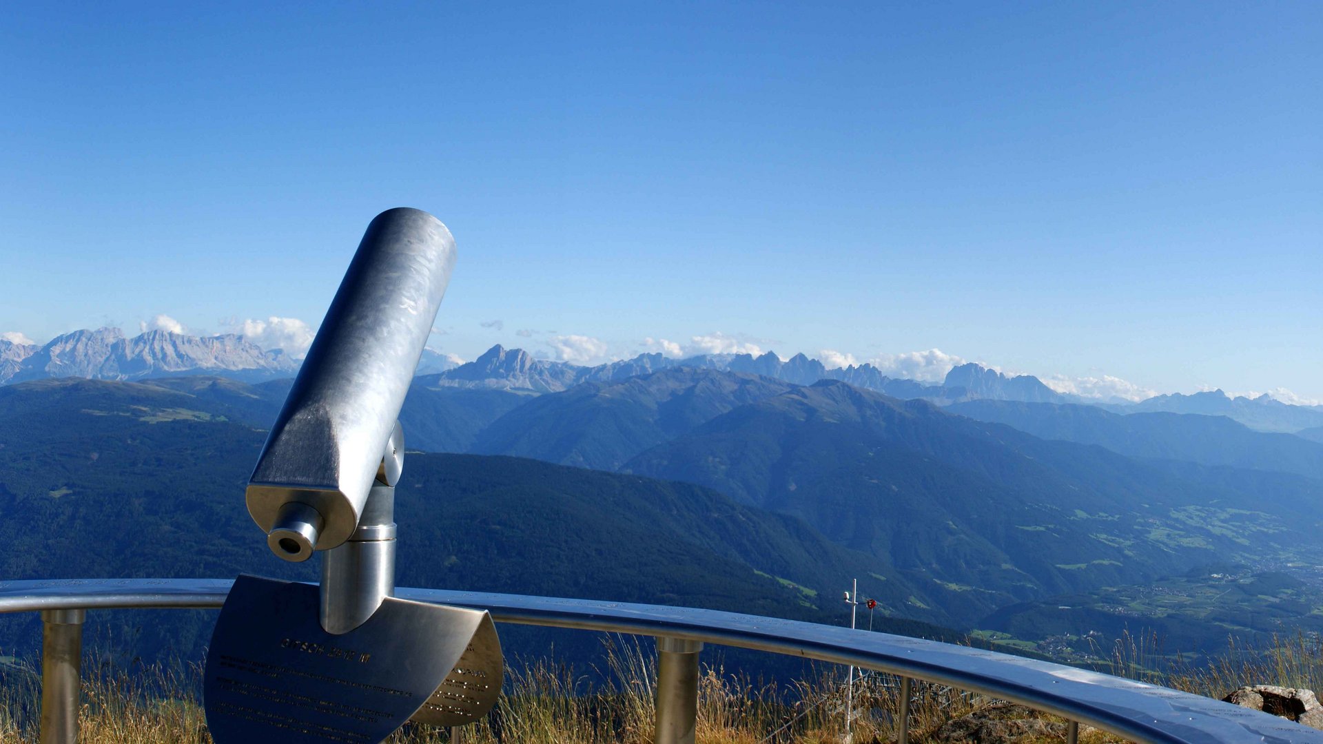 The Gitschberg panoramic platform in Meransen, Italy The picture shows a panoramic view from a mountain summit with a telescope in the center and a summit cross on the right. In the background, vast green valleys and mountains stretch out under a clear blue sky.