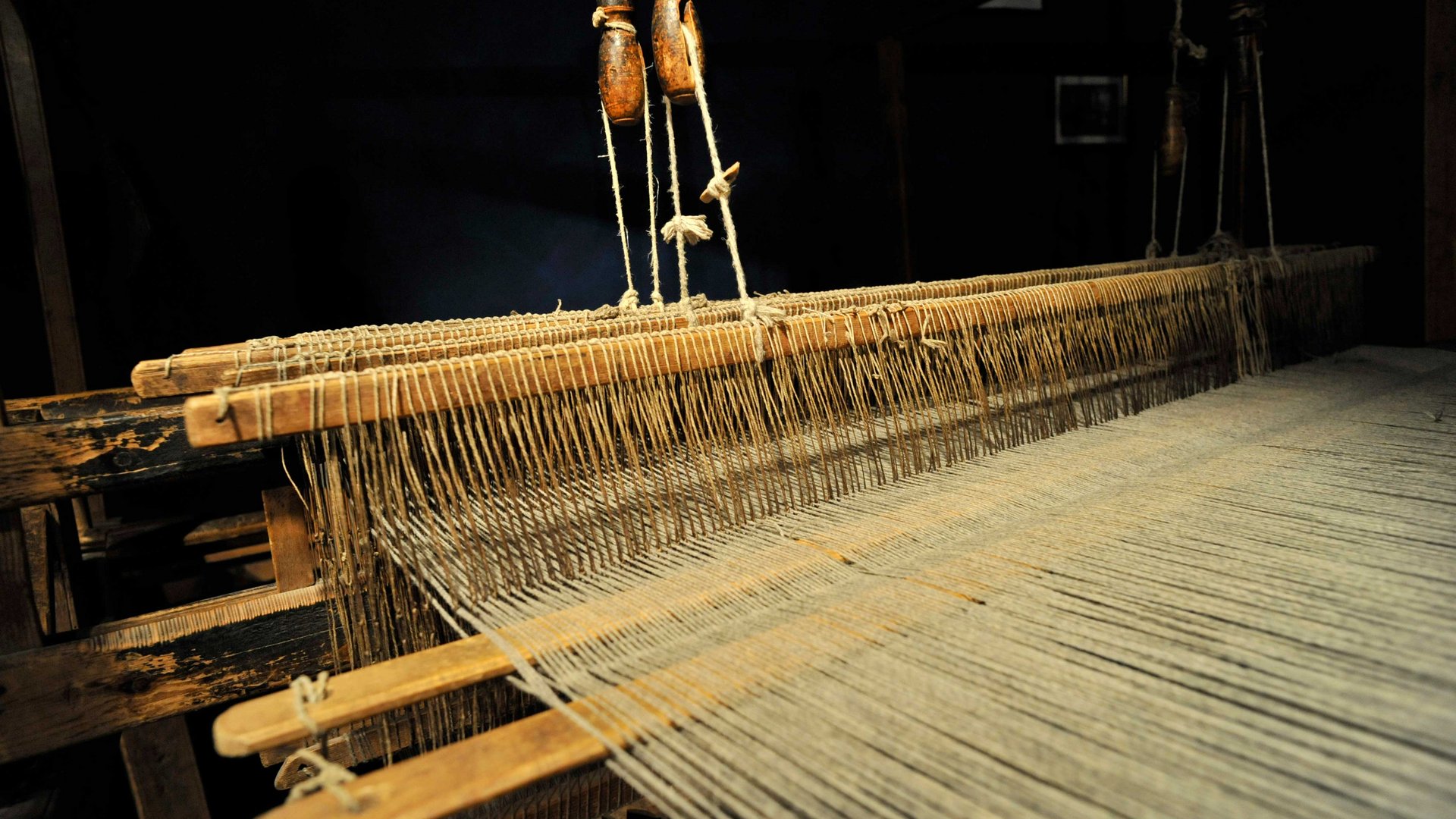 Loden Erlebniswelt in Vintl/Pustertal De afbeelding toont een oude houten weefgetouw waarop draden dicht naast elkaar zijn gespanen. Het weefgetouw wordt in een donkere omgeving tentoongesteld, waardoor de details van de mechaniek en de draden goed zichtbaar zijn.