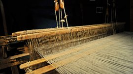 Loden Erlebniswelt in Vintl/Pustertal De afbeelding toont een oude houten weefgetouw waarop draden dicht naast elkaar zijn gespanen. Het weefgetouw wordt in een donkere omgeving tentoongesteld, waardoor de details van de mechaniek en de draden goed zichtbaar zijn.
