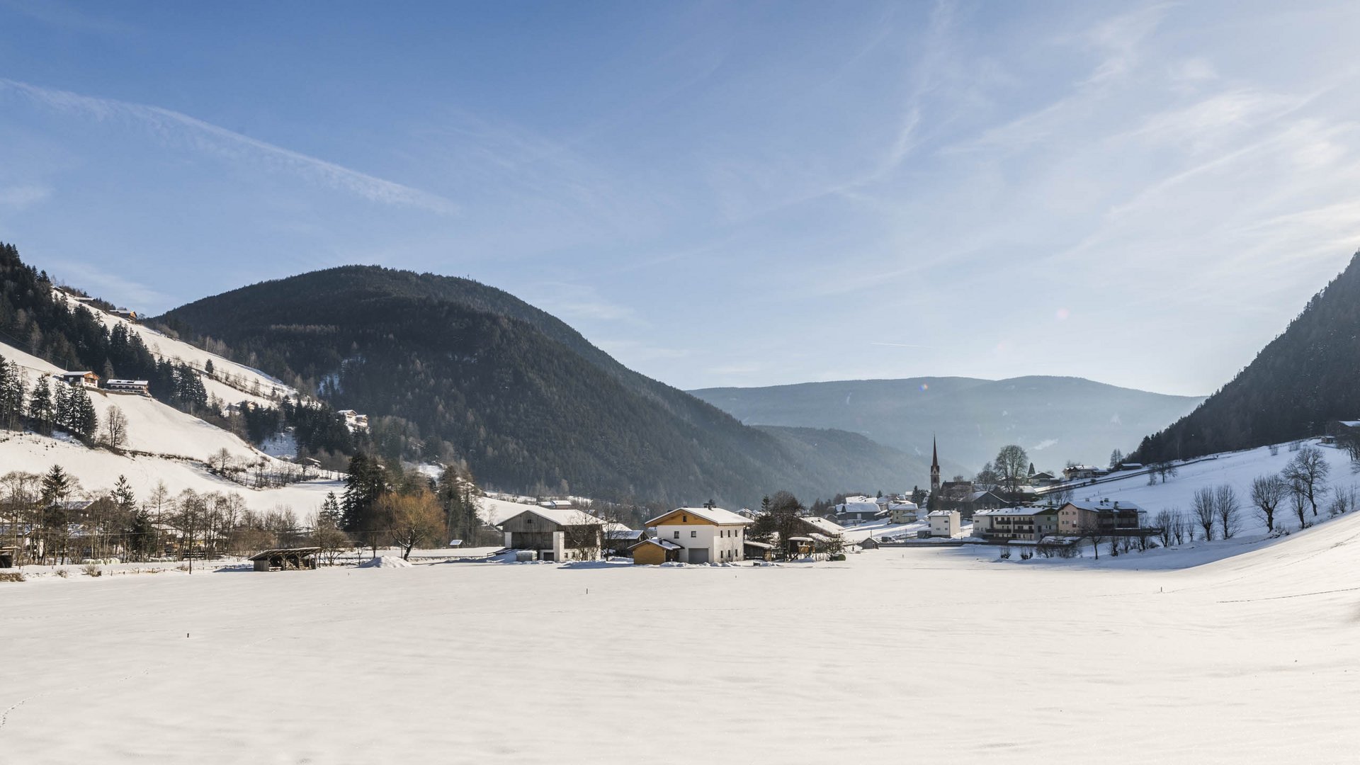 Pfunders: urig und idyllisch Das Bild zeigt ein verschneites Tal mit einem kleinen Dorf in der Mitte, das von schneebedeckten Hügeln und Wäldern umgeben ist. Der Himmel ist klar und blau, und die Sonne scheint hell, wodurch lange Schatten auf den Schnee geworfen werden.