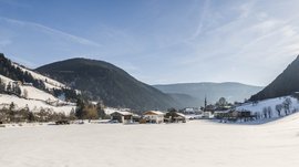 Pfunders: urig und idyllisch Das Bild zeigt ein verschneites Tal mit einem kleinen Dorf in der Mitte, das von schneebedeckten Hügeln und Wäldern umgeben ist. Der Himmel ist klar und blau, und die Sonne scheint hell, wodurch lange Schatten auf den Schnee geworfen werden.