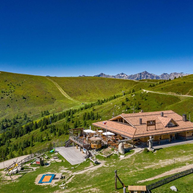 Bergrestaurant Jochtal The picture shows a large mountain hut with an extensive terrace, surrounded by gentle green hills and hiking trails in a rural mountain landscape under a clear blue sky.