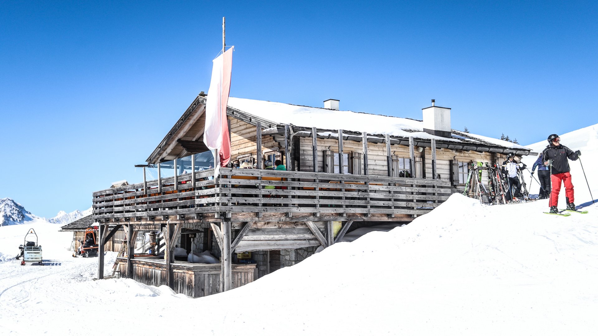 Rund um Meransen: Hütten und Almen Auf dem Bild ist eine rustikale Skihütte aus Holz zu sehen, die von Schnee umgeben ist und auf deren Terrasse einige Gäste sitzen; im Vordergrund steht ein Skifahrer neben abgestellten Skiern.