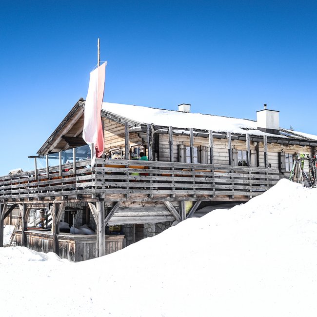 Ochsenalm Auf dem Bild ist eine rustikale Skihütte aus Holz zu sehen, die von Schnee umgeben ist und auf deren Terrasse einige Gäste sitzen; im Vordergrund steht ein Skifahrer neben abgestellten Skiern.