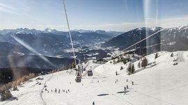 Lyžařská oblast Gitschberg Jochtal Obrázek ukazuje rozsáhlou sjezdovku s mnoha lyžaři sjíždějícími svah. Nad sjezdovkou vede lanovka s gondolami, a v pozadí se rozprostírá impozantní panorama zasněžených hor a údolí pod jasnou modrou oblohou.