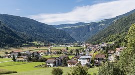 Vintl im Pustertal Das Bild zeigt ein malerisches Dorf in einem grünen Tal, umgeben von bewaldeten Berghängen. In der Mitte des Bildes steht eine Kirche mit einem hohen, spitzen Turm, und das Dorf besteht aus mehreren Wohnhäusern, die sich harmonisch in die Landschaft einfügen.
