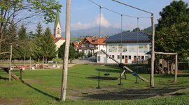 Het Aktivpark Terenten De afbeelding toont een speeltuin met schommels op de voorgrond, gelegen op een groen weiland. Op de achtergrond is een gebouw met zonnepanelen, beschilderde muren en een kerk met een spitse toren te zien, omgeven door bomen en een heuvelachtig landschap.