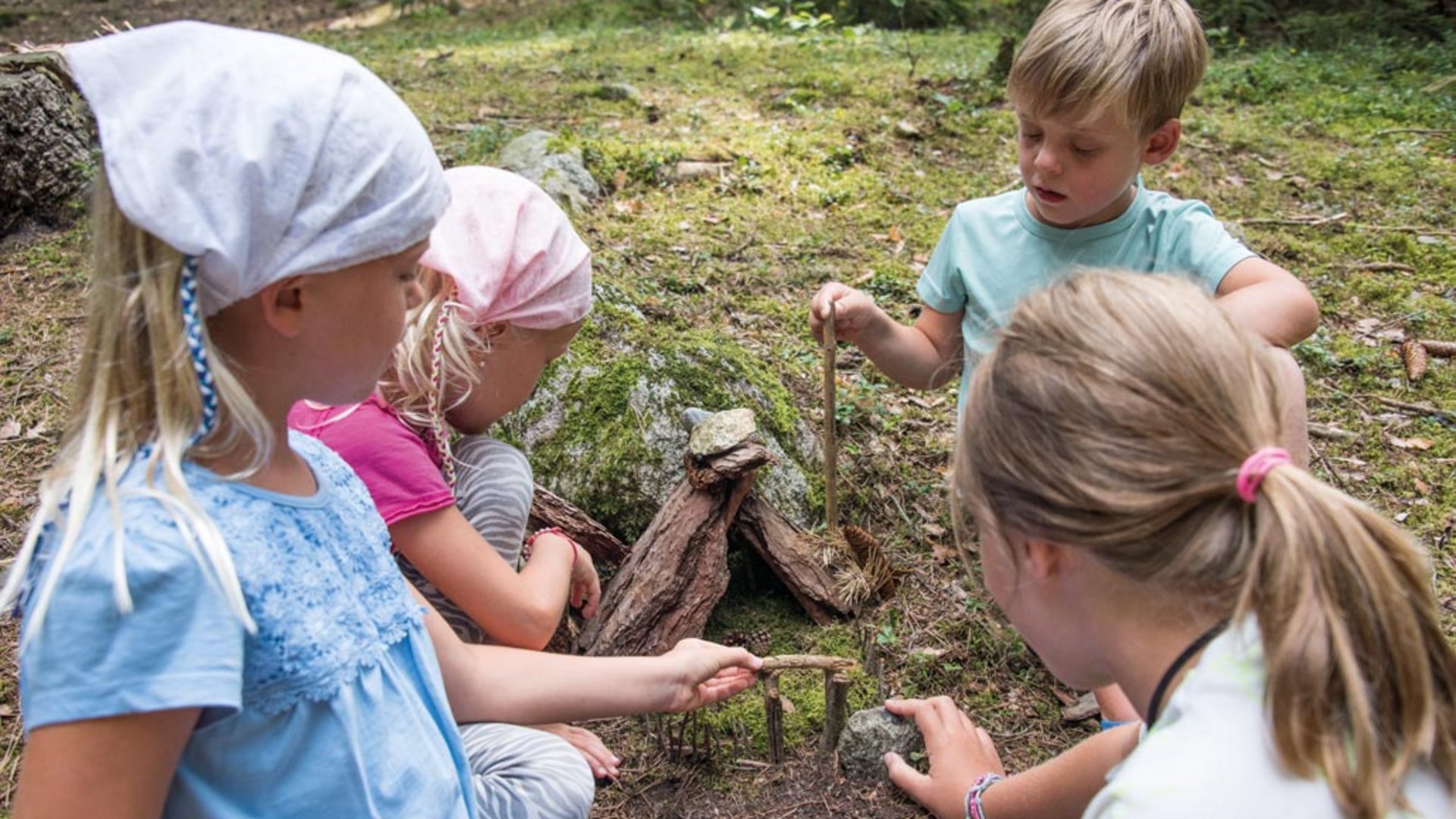 De Elfenweg bij Vintl De foto toont vier kinderen die samen in het bos spelen. Ze bouwen op de grond een kleine structuur van stokjes, stenen en schors, omgeven door mos en bomen.