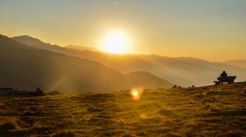 Vals - Jochtal - Steinermandl The picture shows a sunset over a mountain landscape, with the sun low above the mountain peaks casting gentle, golden light onto the meadow. In the foreground, a person sits on a bench looking towards the setting sun.
