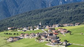 The mountain village of Meransen The picture shows a picturesque village nestled against gentle green hills. In the background, a dense, wooded mountain rises, and a church with a distinctive tower is visible in the center of the village.