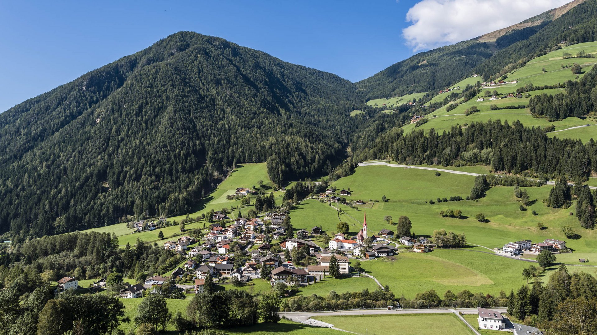 Pfunders: urig und idyllisch Das Bild zeigt eine idyllische, grüne Landschaft mit einem kleinen Dorf, das sich in einem Tal zwischen bewaldeten Berghängen erstreckt. Die Häuser des Dorfes sind von saftigen Wiesen umgeben, und im Hintergrund erhebt sich ein dicht bewaldeter Berg unter einem blauen Himmel mit ein paar weißen Wolken.