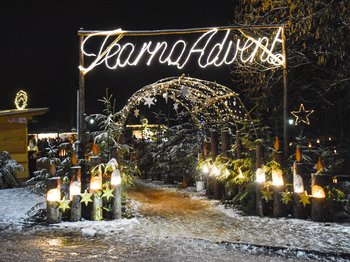 Terenten im Pustertal Das Bild zeigt einen weihnachtlich geschmückten Eingang mit einem großen leuchtenden Schild, auf dem "Tearna Advent" steht. Im Hintergrund sind weitere Lichterketten und Dekorationen, darunter ein leuchtender Stern und kleine Tannenbäume, in einer verschneiten Umgebung zu sehen.