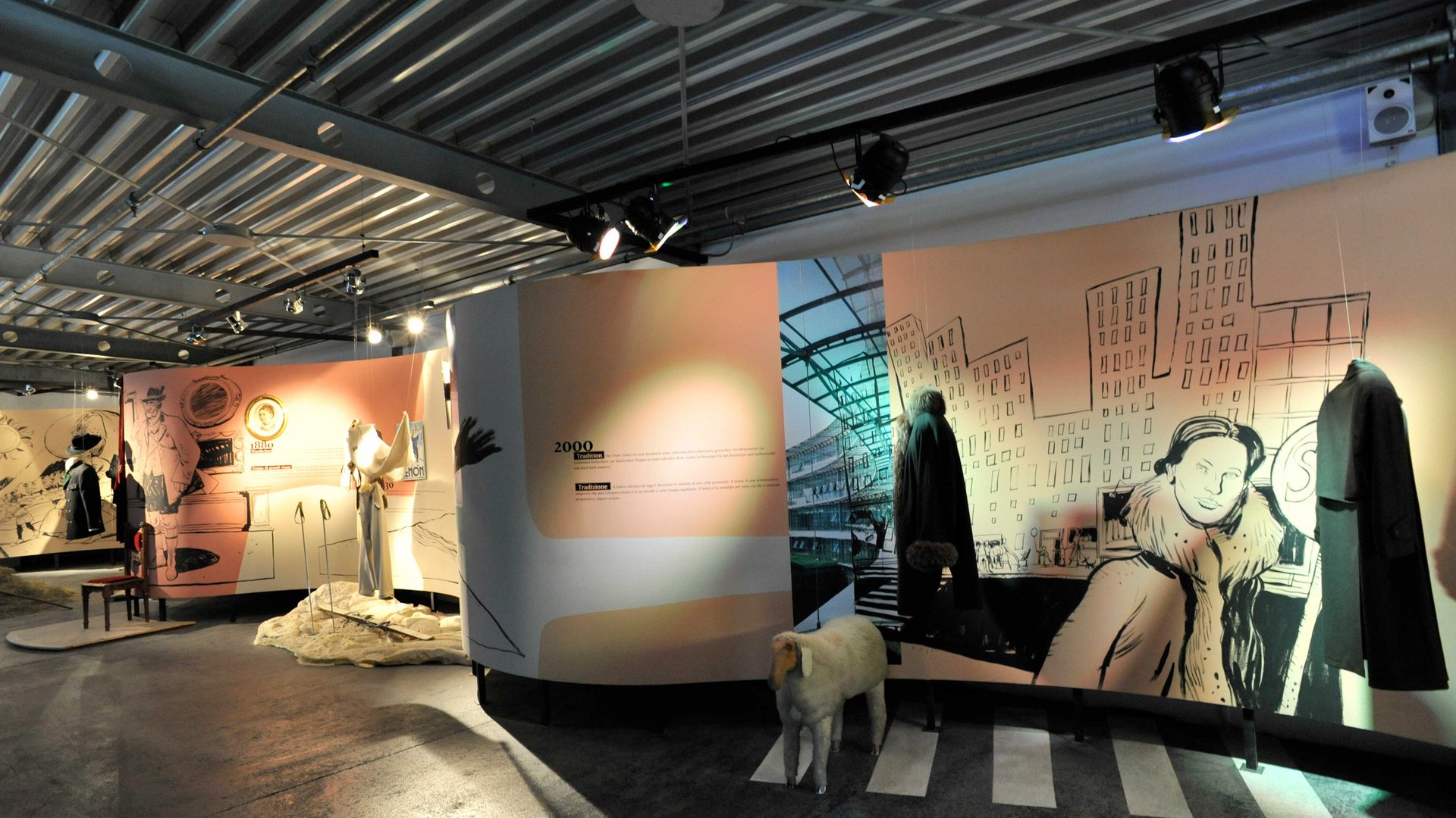 Loden Erlebniswelt in Vintl/Pustertal De afbeelding toont een museuminstallatie waarin historische kleding wordt tentoongesteld. Aan de muren zijn illustraties te zien van taferelen uit verschillende tijdperken, waaronder een stadsgezicht. Voor de muren staan etalagepoppen die jassen en andere kledingstukken dragen, evenals een model van een schaap op de vloer.