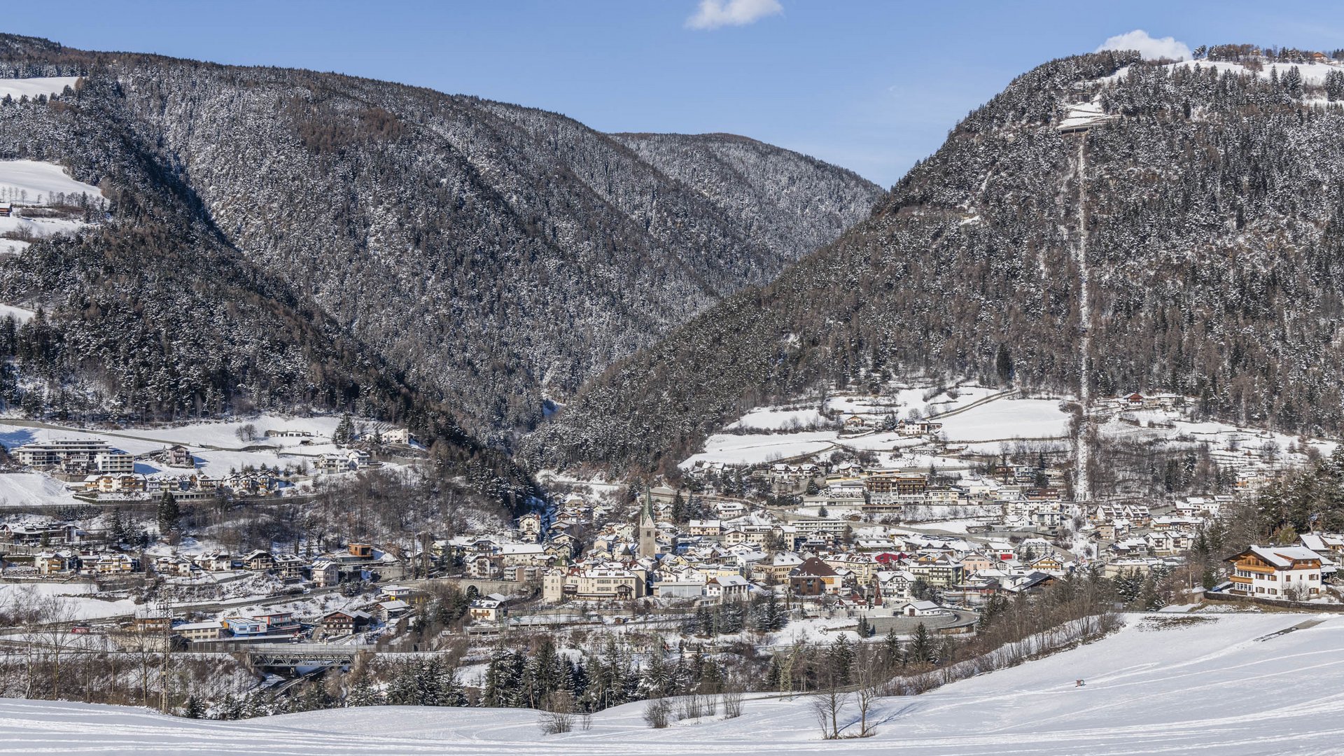 Mühlbach im Pustertal Das Bild zeigt eine Stadt, die in einem schneebedeckten Tal liegt, umgeben von dicht bewaldeten Hügeln. Die Gebäude sind mit Schnee bedeckt und im Hintergrund sind verschneite Hügel und ein klarer blauer Himmel zu sehen.