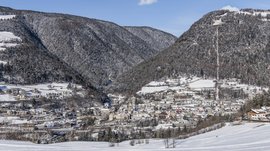 Mühlbach im Pustertal Das Bild zeigt eine Stadt, die in einem schneebedeckten Tal liegt, umgeben von dicht bewaldeten Hügeln. Die Gebäude sind mit Schnee bedeckt und im Hintergrund sind verschneite Hügel und ein klarer blauer Himmel zu sehen.