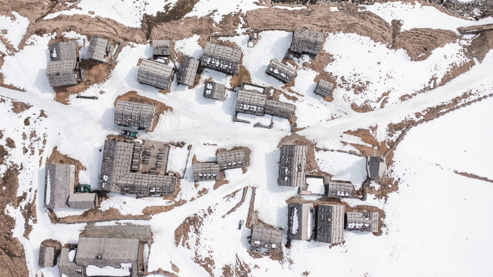 Fane Alm – South Tyrol’s most idyllic Alpine village The image shows a bird's-eye view of a small mountain village with snow-covered wooden roofs. Between the buildings, snowy paths and some exposed brown areas are visible, dominating the winter landscape.
