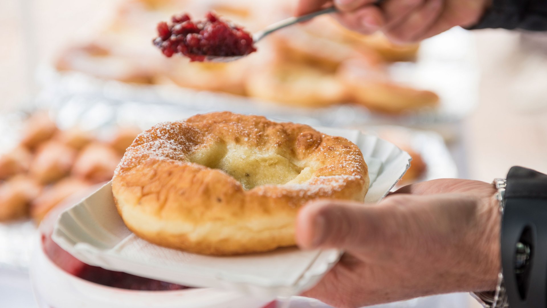 Winkelen in Mühlbach/Italië De afbeelding toont een persoon die met een lepel rode jam of compote op een gefrituurd gebakje schept, dat met de hand op een papieren bordje wordt gehouden. Het gebakje is bestrooid met poedersuiker en ziet er goudbruin en knapperig uit.