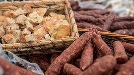 Winkelen in Mühlbach/Italië De afbeelding toont een mand met stukken brood, die rustiek gesneden zijn. Naast de mand liggen talrijke kruidige, luchtgedroogde worsten in een andere gevlochten doos. De scène wijst op een traditionele markt- of boerderijomgeving.