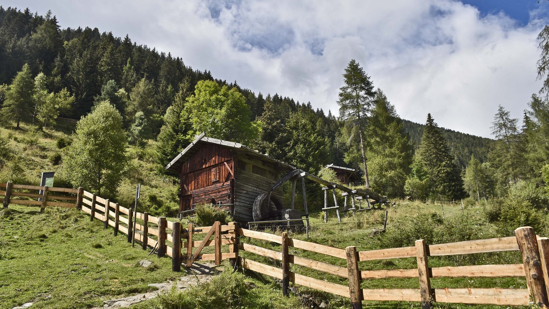 Der Mühlenweg in Terenten Holzhaus mit Wasserrad auf grüner Wiese vor Wald und blauem Himmel