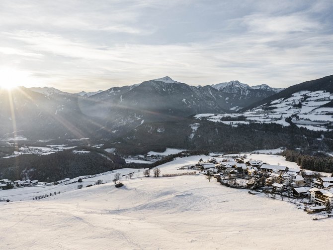 The Alpine hamlet of Spinges The picture shows a snowy landscape in the mountains, shortly after sunrise. The sun rises behind the mountains and shines light over the valley, which is characterized by snow-covered fields and hills. On the right side of the picture there is a small village, also covered with snow. The mountains in the background are partially forested and visible in the distance.