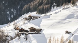 Pfunders: urig und idyllisch Das Bild zeigt eine winterliche Berglandschaft mit schneebedeckten Hügeln und mehreren Berghütten, die an den Hängen verteilt sind. Im Hintergrund ist ein dichter Wald zu sehen, und das Licht der tief stehenden Sonne wirft lange Schatten auf den Schnee.
