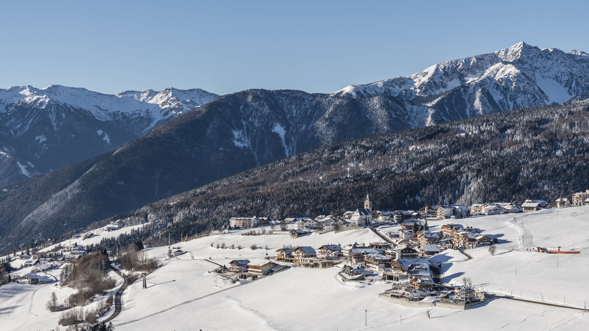 The mountain village of Meransen The picture shows a snowy village in a valley, surrounded by forested mountain slopes and snow-covered peaks. A church with a tower is centrally placed in the village, and a road winds through the white landscape. The sky is clear and blue, creating a sunny winter atmosphere.