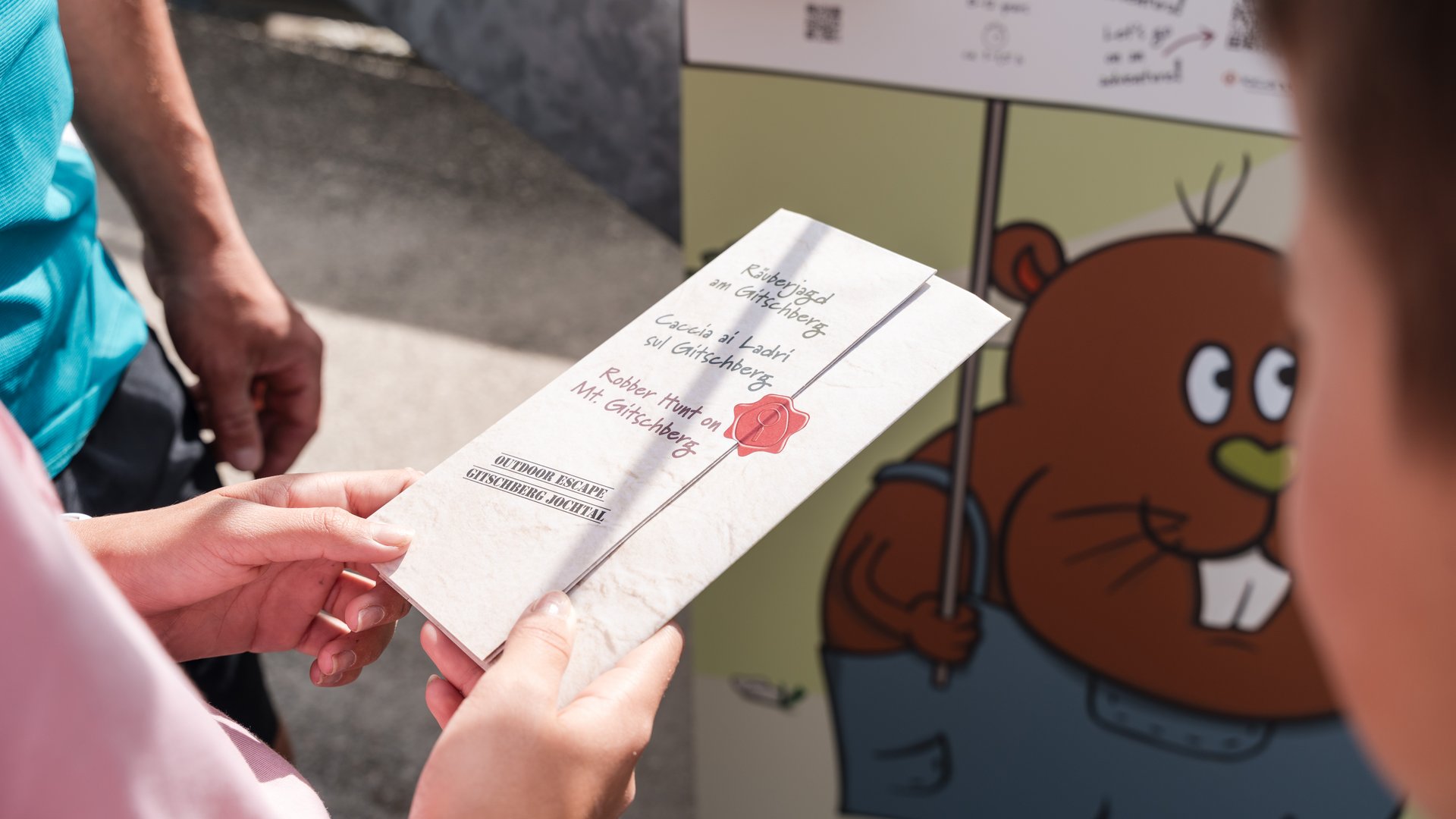 Outdoor Escape on Mt. Gitschberg Person holding flyer for Robber Hunt on Mt. Gitschberg in front of colorful board