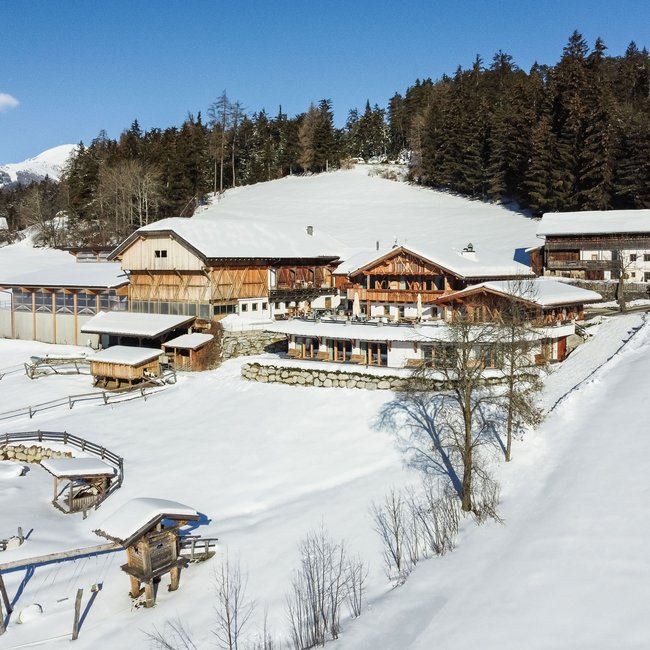 Ahner Berghof Obrázek ukazuje tradiční alpské dřevěné domy, obklopené zasněženou krajinou s jasně viditelnou turistickou stezkou, před zalesněným pozadím a pod modrou oblohou.