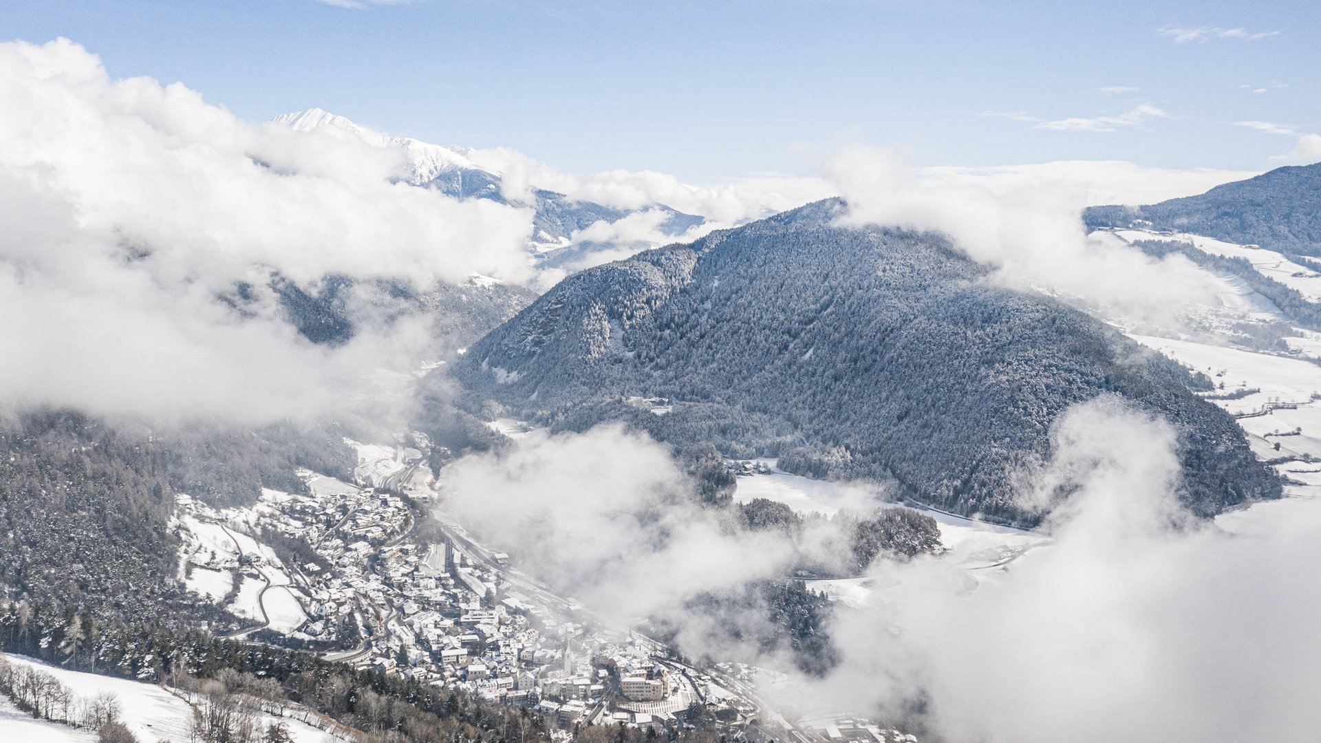 Mühlbach im Pustertal Das Bild zeigt eine verschneite Landschaft mit einem Tal und einer kleinen Stadt, die von bewaldeten Hügeln und Bergen umgeben ist. Wolken ziehen über die Szenerie, wodurch Teile der Landschaft verdeckt werden und ein malerischer, winterlicher Eindruck entsteht.