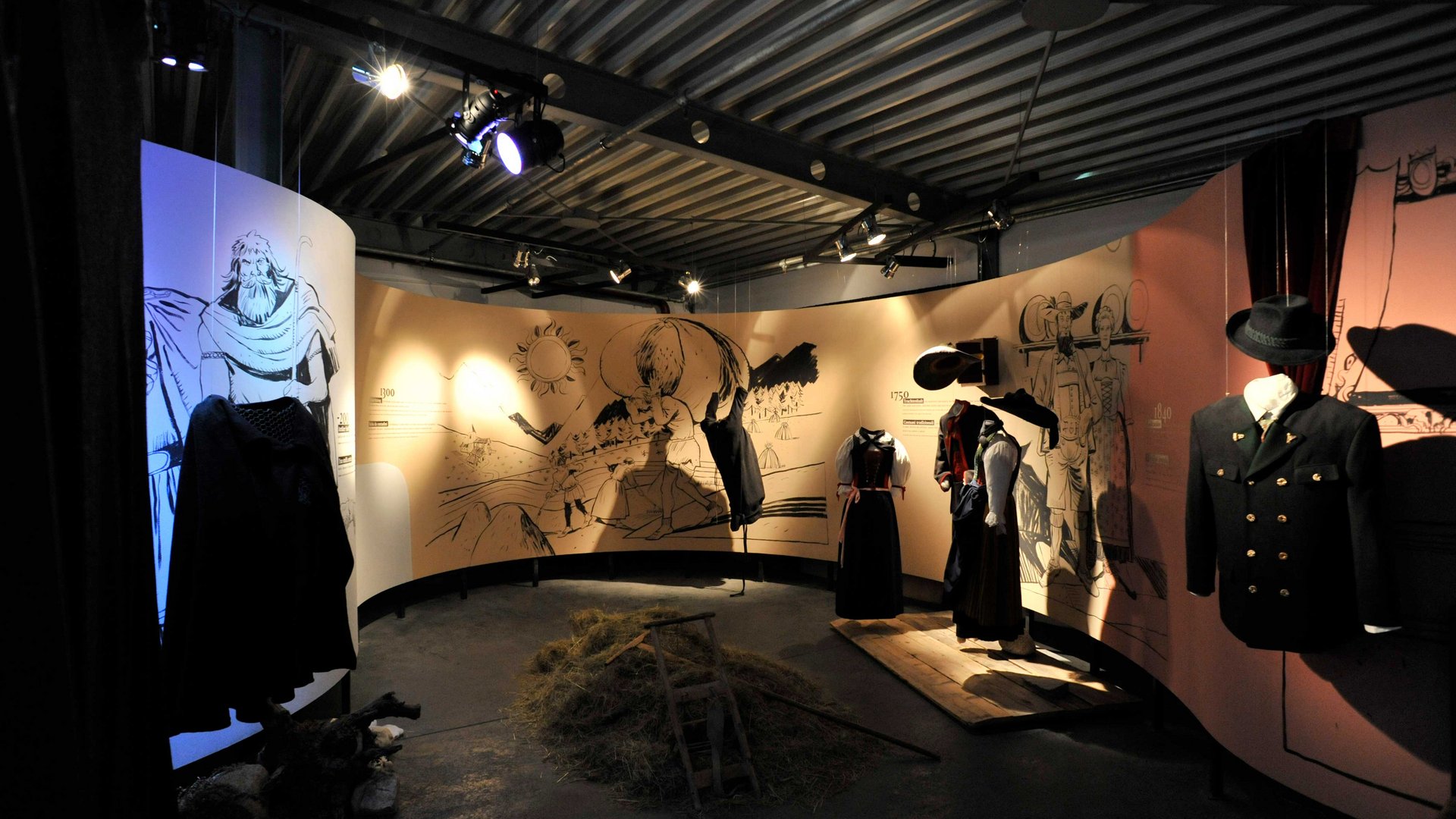 Loden Erlebniswelt in Vintl/Pustertal De afbeelding toont een tentoonstelling met historische kledingstukken die op etalagepoppen worden gepresenteerd. Op de achtergrond zijn grote geïllustreerde muurschilderingen met historische taferelen en teksten te zien die de context van de tentoongestelde kleding uitleggen.