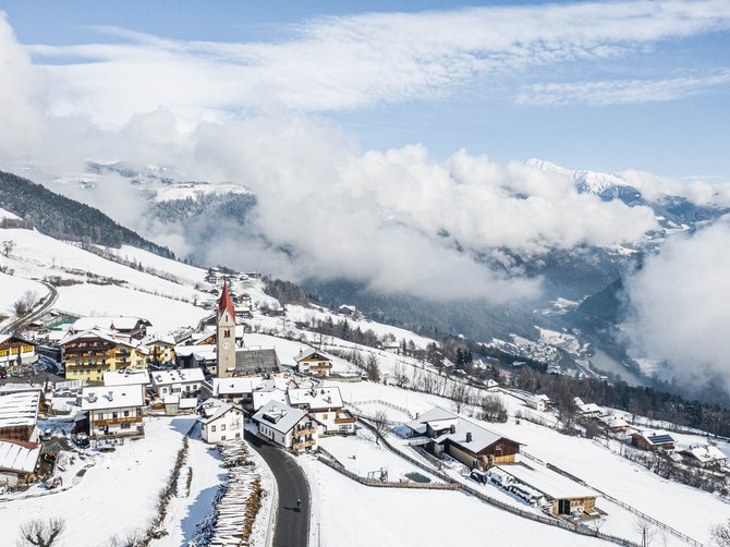 The Alpine hamlet of Spinges The picture shows a snowy mountain village with a church whose red tower stands out among the snow-covered houses. In the background, you can see wooded hills and low-hanging clouds that partially obscure the valley below.