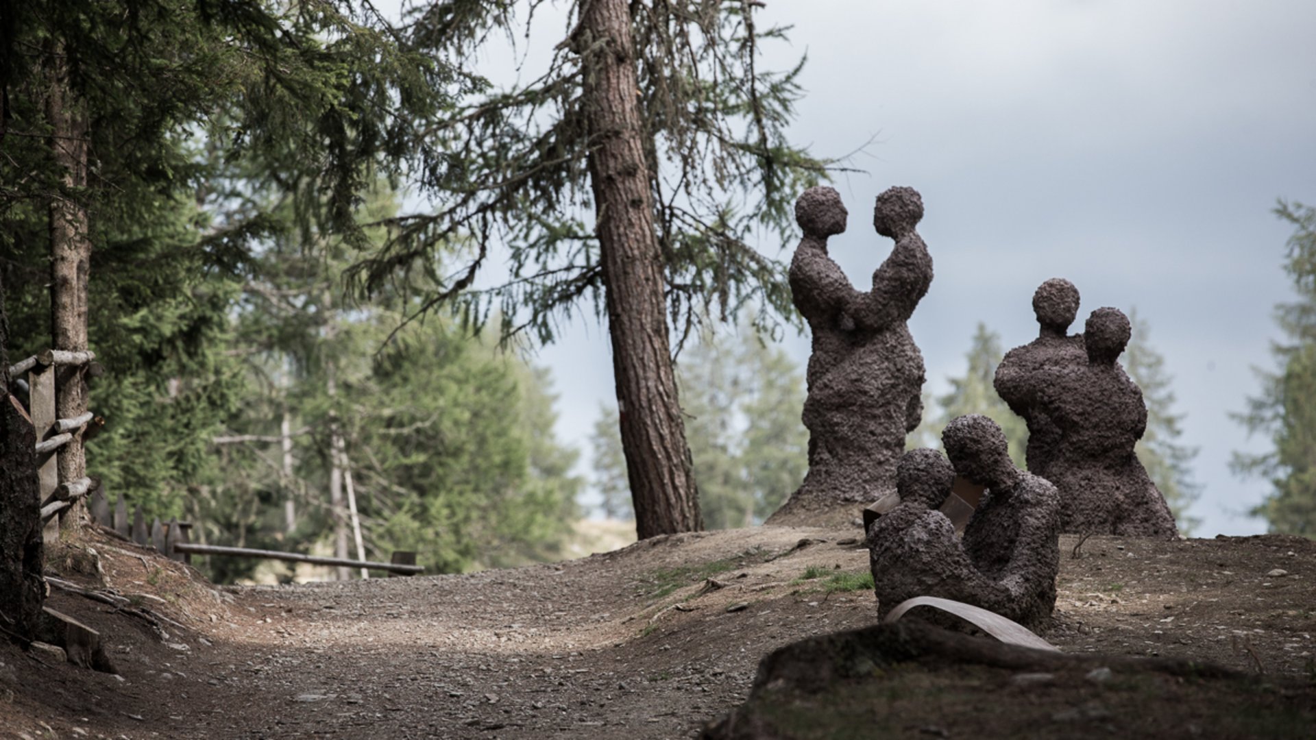 Der Schöpfungsweg in Rodeneck Das Bild zeigt mehrere abstrakte Skulpturen von Paaren, die aus grobem Material geformt sind und sich in einem Waldgebiet auf einem Weg befinden. Die Figuren stehen oder sitzen nah beieinander, als ob sie sich umarmen oder in engem Kontakt sind.