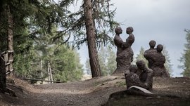 Der Schöpfungsweg in Rodeneck Das Bild zeigt mehrere abstrakte Skulpturen von Paaren, die aus grobem Material geformt sind und sich in einem Waldgebiet auf einem Weg befinden. Die Figuren stehen oder sitzen nah beieinander, als ob sie sich umarmen oder in engem Kontakt sind.