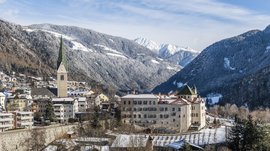 Mühlbach im Pustertal Das Bild zeigt eine verschneite Stadt in einem Tal, umgeben von bewaldeten Bergen. Im Vordergrund ist eine Kirche mit einem hohen, spitzen Turm und mehreren historischen Gebäuden zu sehen, während im Hintergrund schneebedeckte Berge die Szene dominieren.