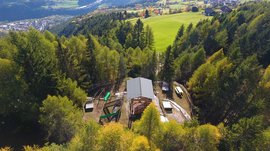 The recreation area in Spinges, South Tyrol The picture shows a forest landscape with a small building in the middle, surrounded by trees. In the background, green hills and valleys extend, with scattered houses and fields visible.