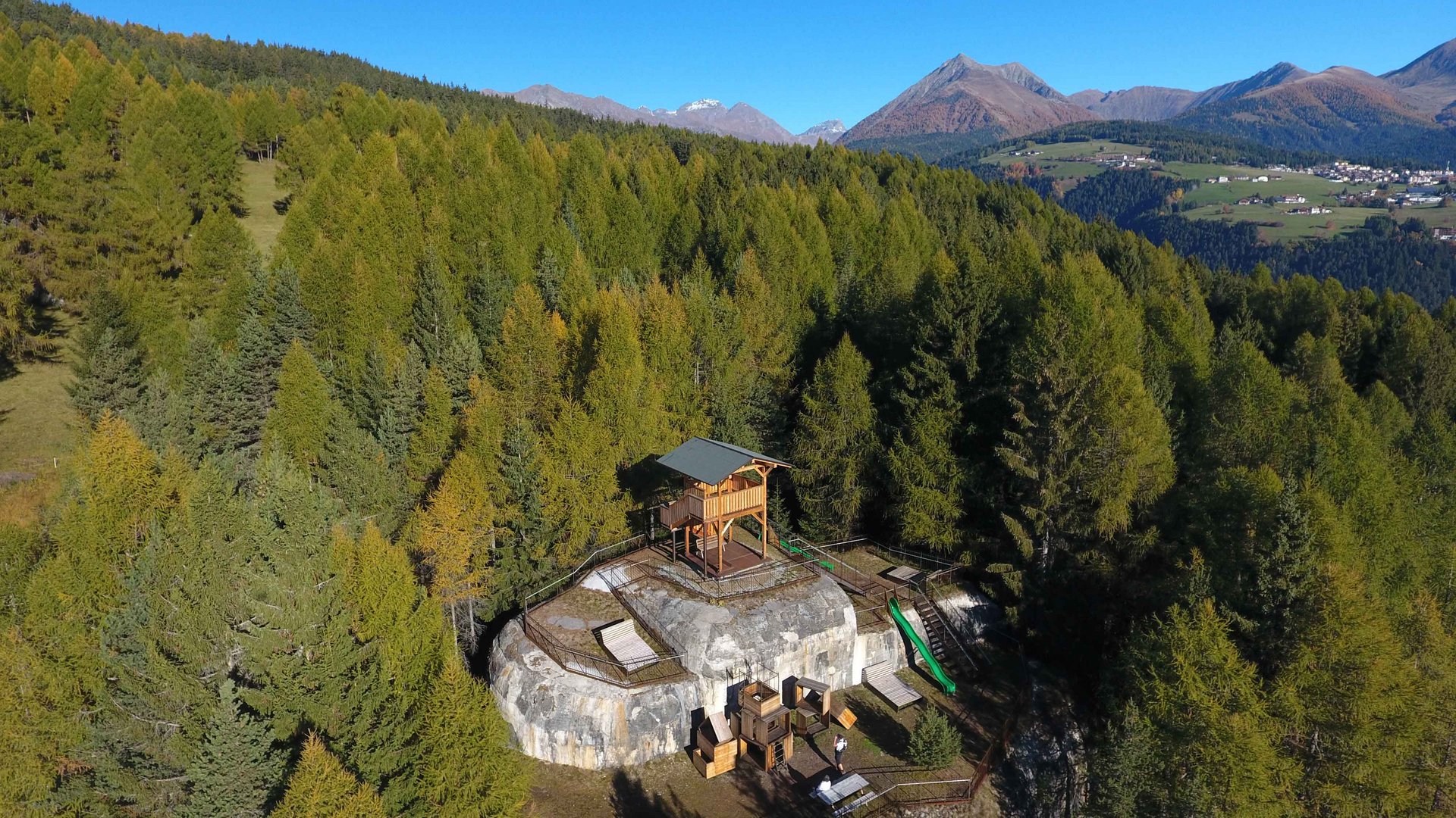 The recreation area in Spinges, South Tyrol The picture shows a wooden observation platform located on a rocky outcrop in the middle of a dense forest. In the background, green mountains and a clear blue sky can be seen, offering a wide view over the landscape.