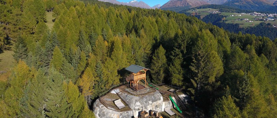 The Gitschberg webcams The picture shows a wooden observation platform located on a rocky outcrop in the middle of a dense forest. In the background, green mountains and a clear blue sky can be seen, offering a wide view over the landscape.