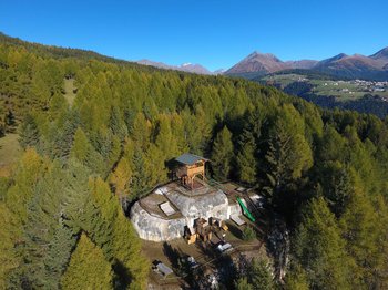 The Alpine hamlet of Spinges The picture shows a wooden viewing platform located on a rocky outcrop in the middle of a dense forest. Green mountains and a clear blue sky can be seen in the background, offering a sweeping view over the landscape.