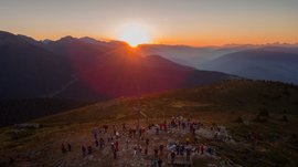 Vals - Jochtal - Steinermandl The image shows a sunset over a mountain landscape, with numerous people gathered on a mountain peak. In the center of the image stands a large summit cross, while the sky and mountains glow in warm orange and red tones.