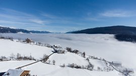 The mountain village of Meransen The picture shows a winter mountain landscape with snow-covered fields and scattered houses on a hill. In the background, a dense sea of clouds stretches over the valley, while the snow-capped mountain peaks emerge under a clear, blue sky.