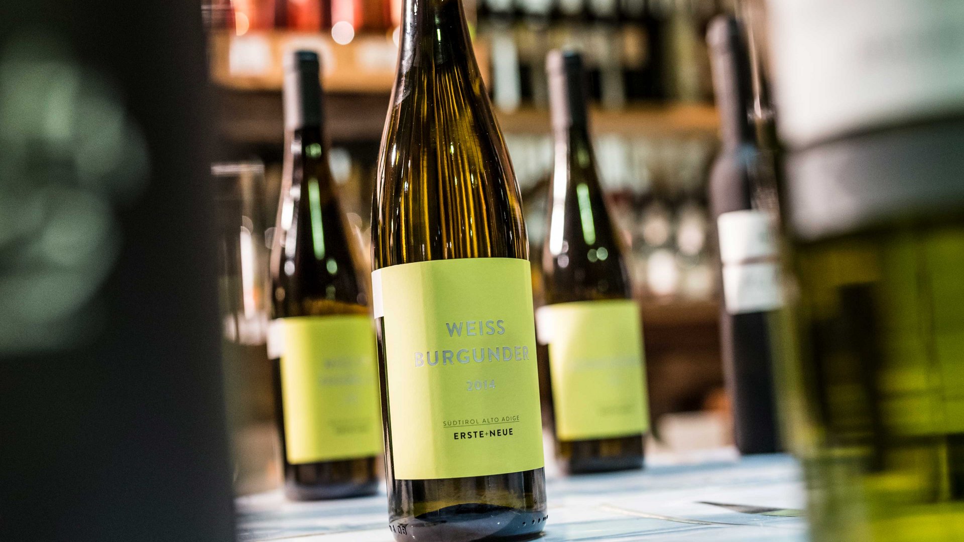 Winkelen in Mühlbach/Italië De afbeelding toont een fles Weißburgunder-wijn met een geel etiket waarop "Weiss Burgunder 2014" staat. Op de achtergrond zijn andere wijnflessen vaag te zien, die ook op een plank staan.