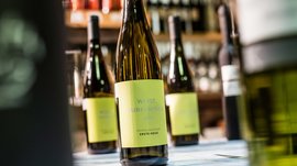 Winkelen in Mühlbach/Italië De afbeelding toont een fles Weißburgunder-wijn met een geel etiket waarop "Weiss Burgunder 2014" staat. Op de achtergrond zijn andere wijnflessen vaag te zien, die ook op een plank staan.