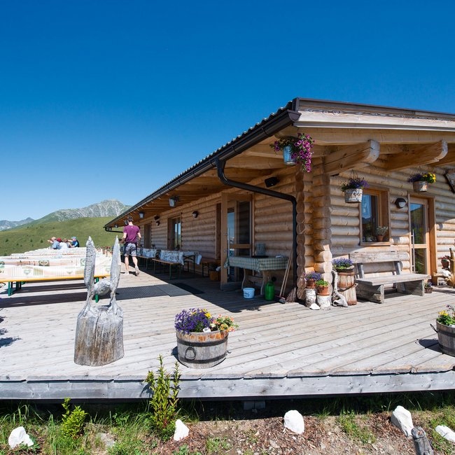 Nesselhütte De afbeelding toont een gezellig houten berghutje met een brede veranda waarop tafels en banken staan. Het hutje is omgeven door bloeiende bloemen en biedt een wijds uitzicht op groene heuvels en bergen onder een stralend blauwe lucht.