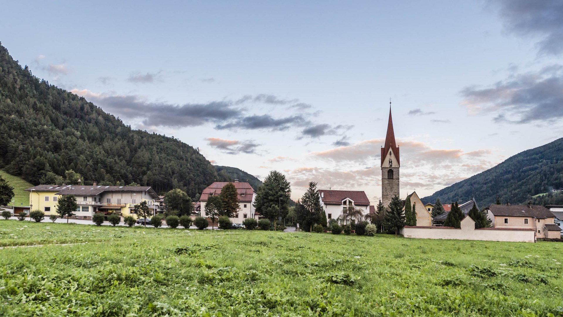 Vintl im Pustertal Das Bild zeigt eine grüne Wiese im Vordergrund mit einer Kirche und mehreren Häusern im Hintergrund. Die Kirche hat einen markanten roten Kirchturm, und das Dorf ist von bewaldeten Hügeln umgeben, während der Himmel leicht bewölkt ist.
